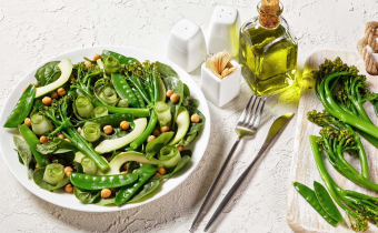 Green vegetable salad of sauteed tenderstem broccoli, snow peas, avocado, cucumber, chickpeas, baby spinach with olive oil dressing, served on a white plate with cutlery on a white table, top view