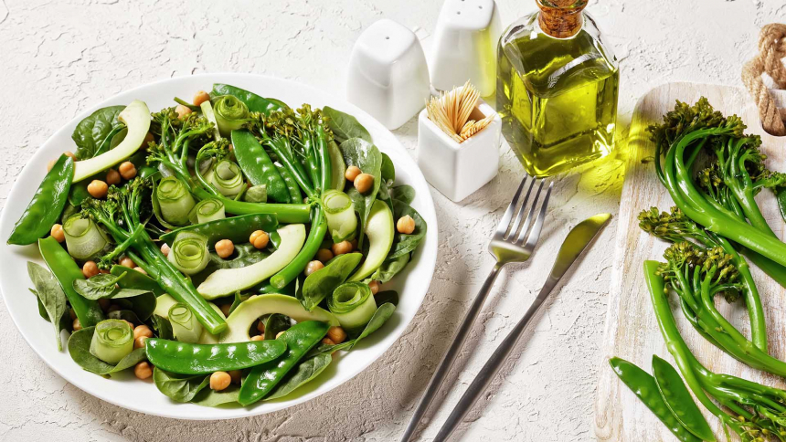Green vegetable salad of sauteed tenderstem broccoli, snow peas, avocado, cucumber, chickpeas, baby spinach with olive oil dressing, served on a white plate with cutlery on a white table, top view